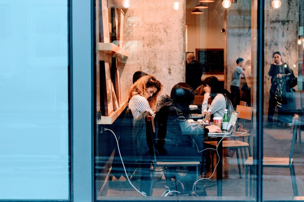 An office with laptops and books through a glass door showing people working on devices